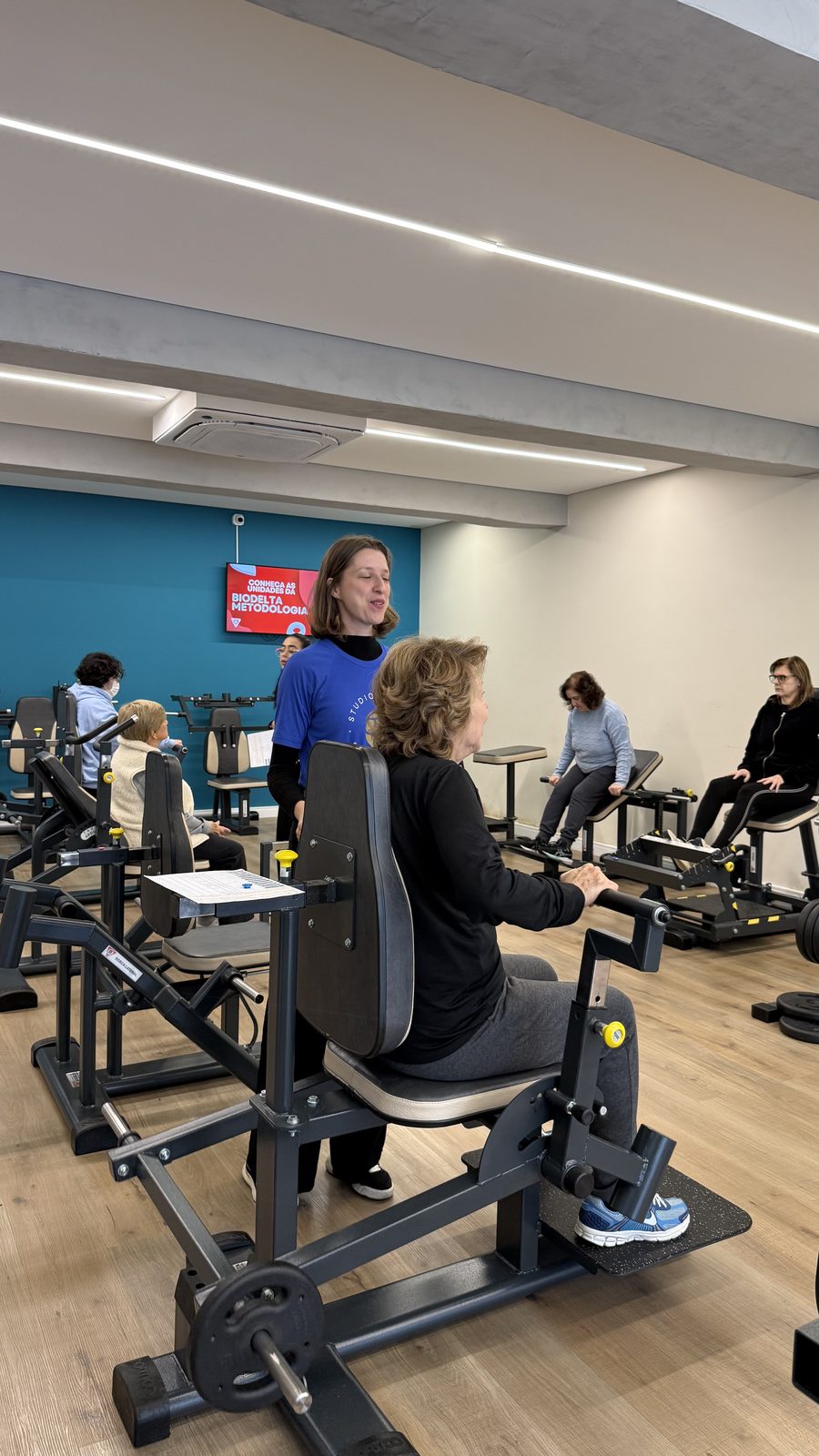 Members training on strength machines on the Studio Tekoá floor while a professional guides a seated member